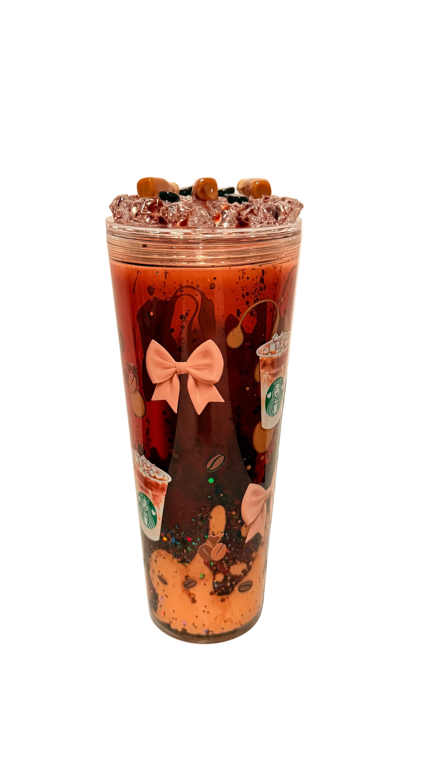 A clear acrylic tumbler cup filled with a layered drink, featuring a dark brown top layer, an orange-brown middle layer, and a light orange bottom layer. The cup is decorated with a pink bow graphic and coffee cup illustrations, topped with crushed ice and small chocolate pieces.